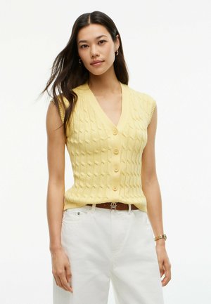 Donna con lunghi capelli scuri che indossa un gilet di maglia giallo senza maniche con bottoni e pantaloni bianchi con cintura marrone.