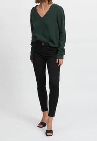 Ciemnozielony sweter prążkowany z dekoltem w kształcie litery V, zestawiony z czarnymi dopasowanymi dżinsami i czarnymi klapkami. Gładka faktura i swobodny styl.