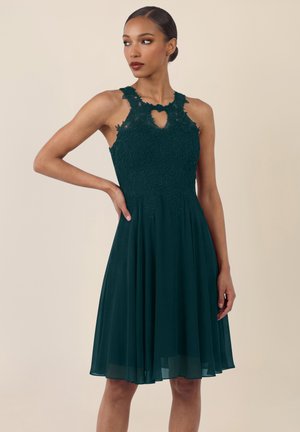 Kvinde i en ærmeløs mørkegrøn knælængde kjole med blonder på overdelen og nøglehulsudskæring, stående med den ene hånd på hoften.