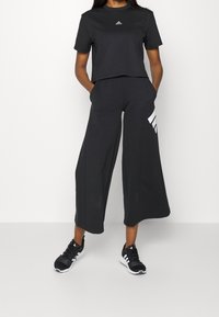 Svart kort t-shirt med en liten vit Adidas-logotyp, kombinerad med vida svarta byxor med vita randiga detaljer. Svarta sportskor.