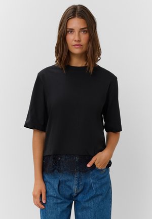 Jeune femme aux cheveux bruns mi-longs porte un haut noir ample avec bordure en dentelle et un jean bleu, debout devant un fond clair uni.
