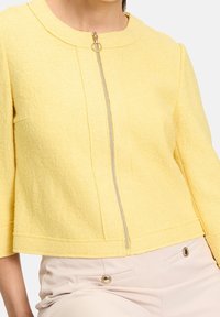 Žlutá zkrácená bunda z texturovaného materiálu, s předním zipem a kulatým výstřihem. Má tříčtvrteční rukávy a minimální detaily.