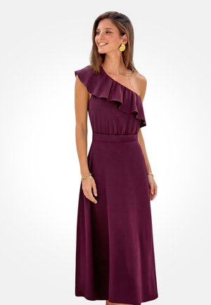 Frau lächelnd, trägt ein ärmelloses Pflaumenkleid mit einem gerafften, einseitigen Ausschnitt, goldene Ohrringe und goldene Armbänder, stehend vor einem weißen Hintergrund.