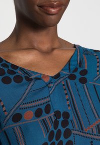 Blå mönstrad plagg med svarta och bruna cirkulära och linjära mönster, med V-ringning och knapprad vid bröstet.