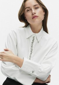 Biała koszula z kołnierzykiem, zdobiona dekoracyjnymi, srebrnymi koralikami na przodzie oraz z guzikami w mankietach z dopasowanymi akcentami. Gładka faktura.