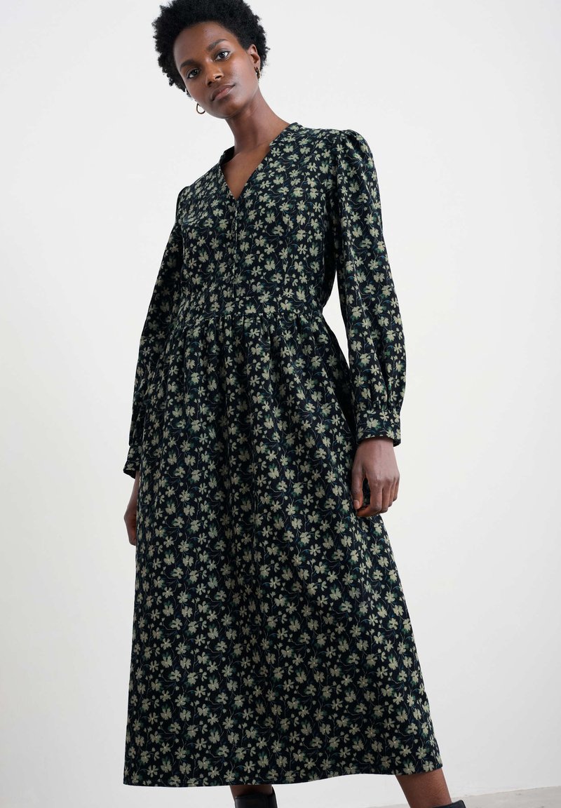 Langarm, v-hals jurk van donkere stof met een bloemenprint in crème en groen. Heeft een taillerende taille en een uitlopende rok. Zachte textuur.