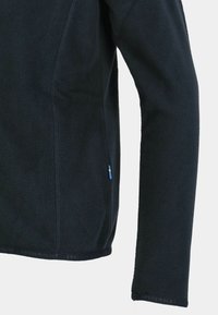 Musta fleece-takki, jossa on tekstuuri, pitkät hihat ja pieni sininen ja keltainen etiketti helman lähellä. Brändin nimi näkyy alhaalla.