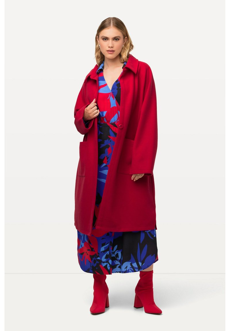 Manteau en laine rouge au fit oversized, avec un col, deux poches avant et un bouton unique. En dessous se trouve une robe à motifs floraux en bleu et rouge.