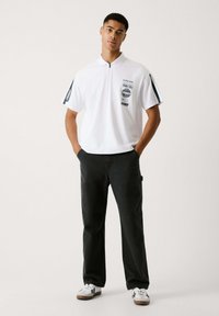 Camisa polo blanca con cremallera frontal, detalles en azul marino y logotipos impresos; combinada con pantalones oscuros y zapatillas blancas con suelas marrones.