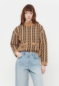 Strikkegenser i kamelfarge med svart kabelstrikkmønster, høy krage, to knappede frontlommer, kombinert med høyt midje lyseblå jeans.