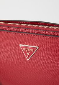 Rød tekstureret pung med en gylden lynlås, der har et trekantet logo-tag med teksten "GUESS USA." Glat overflade med synlige syningsdetaljer.