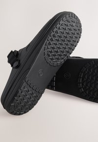 Chaussures noires avec une semelle en caoutchouc texturée, dotées d'une sangle pour un ajustement sécurisé. Matériau supérieur lisse avec des détails perforés.