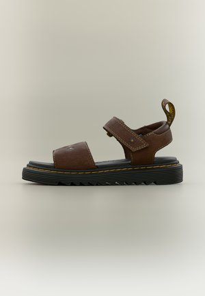 Καφέ δερμάτινο σανδάλι με ρυθμιζόμενους ιμάντες, μαύρη μαλακή σόλα, κίτρινη ραφή και θηλιά στη φτέρνα σε ουδέτερο φόντο.