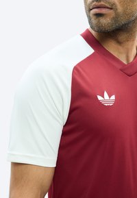 Mies, joka käyttää tummanpunaista Adidas-paitaa valkoisilla hihoilla, jossa on rinnan kohdalla logo ja V-pääntie.