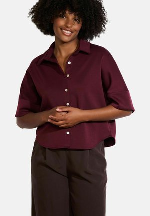 Blusa colletto corto color borgogna con orlo arrotondato e chiusura a bottoni. Tessuto morbido e leggero. Abbinata a pantaloni marrone scuro.