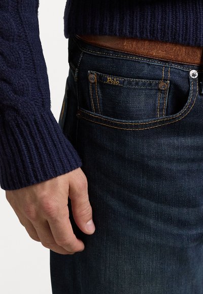 La manica del maglione a trecce blu navy incontra i jeans in denim blu scuro. I jeans presentano una toppa in pelle beige e cuciture gialle, con dettagli delle tasche ben visibili.
