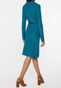 Turkos klänning med strukturerade vertikala ränder, långa ärmar och en åtsittande midja. Buren med bruna klacksandaler, visad bakifrån.