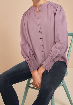 Fialová košile s knoflíky, se žabičkovanými rukávy, límečkem a pěti knoflíky vpředu. Měkká látka s hladkým povrchem.