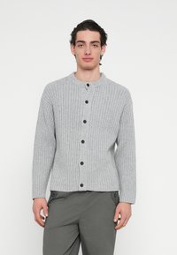 Grå strikket cardigan med ribbet struktur, rund halslinning og knappelukking foran. Har lange ermer og kontrasterende svarte knapper.