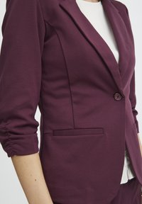 Vinröd skräddarsydd blazer med en slät yta, enkelknapplåsning och strukturerade slag. Har en sidoficka och detaljer med samlad ärm.