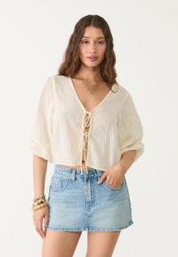 Kremno obarvana, lahka krajša bluza z V-izrezom in zavezanjem, v kombinaciji z modrim denim mini krilom. Zlata ogrlica dopolnjuje videz.
