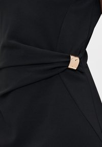 En svart kjole med vridd stoffdetalj i midjen, med en metallisk rosé gullspenne for ekstra detaljer og struktur.