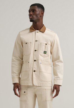 Beige denim jakk, millel on pruun krae, kahel rinnal taskul, kontrastsete õmblustega ja logo detailiga.