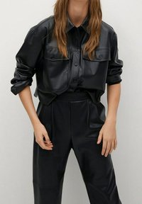 Camisa de cuero negro con botones, dos bolsillos en el pecho, mangas enrolladas y un corte relajado, combinada con pantalones de cuero negro.