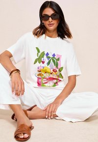 T-shirt blanc présentant un graphisme coloré de fruits, de plantes et de vaisselle. Le modèle porte un pantalon clair et des sandales marron, avec des lunettes de soleil et des accessoires.