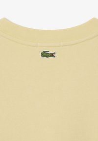 Sudadera de algodón amarilla con cuello acanalado y un pequeño logo de cocodrilo verde bordado en la parte trasera. Textura suave y diseño casual.