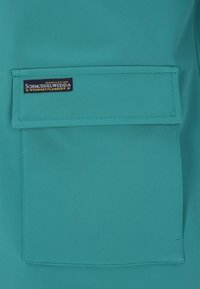 Țesătură verde-albăstrui cu textură netedă, prevăzută cu un buzunar orizontal aplicat. O etichetă albastră cusută pe buzunar afișează numele brandului "Schmuddelwedda."