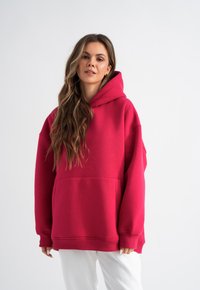 Felpa oversize fucsia con una grande tasca frontale, polsini a costine e cappuccio con cordino. Il tessuto sembra morbido e liscio.