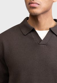 Polo shirt tricotat de culoare maro închis, cu o textură ribbată, având guler în V și ușor întors, purtat deasupra unei cămăși cu guler rotund de culoare albă.