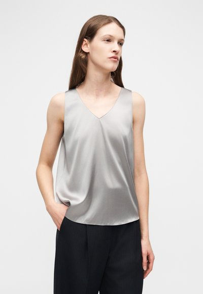 Femme portant un haut sans manches en satin argenté à encolure en V et un pantalon noir, debout, une main dans la poche, sur un fond clair uni.