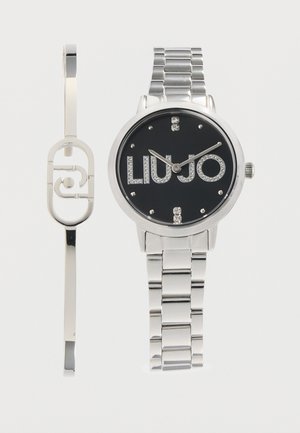 Sølv metal ur med sort urskive og krystaller på "LIU JO" logoen, kombineret med et matchende sølv armbånd med ovalt hulmønster.