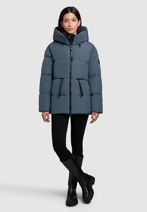 Pufferjack in leisteenblauw met een hoge kraag, zwarte accenten, twee voorzakken en een ritssluiting. Het model draagt zwarte leggings en laarzen tot de knie.