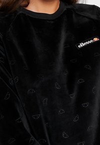 Sudadera de terciopelo negra con sutiles patrones semicirculares en relieve y un pequeño parche blanco con el logo "ellesse" en el pecho.