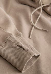 Hoodie beige avec des poignets côtelés et une finition texturée. Cordon de serrage au cou et un tissu doux et lisse. Détails de couture subtils.