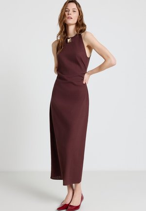 Donna in piedi che indossa un vestito senza maniche di colore marrone scuro, lungo fino alla caviglia, con fibbia quadrata e ballerine rosse con punta, su uno sfondo semplice.