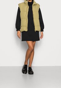 Doudoune sans manches vert olive, fermeture éclair, avec un col haut. Robe à manches longues noire en dessous. Le modèle porte des chaussures noires vernies.