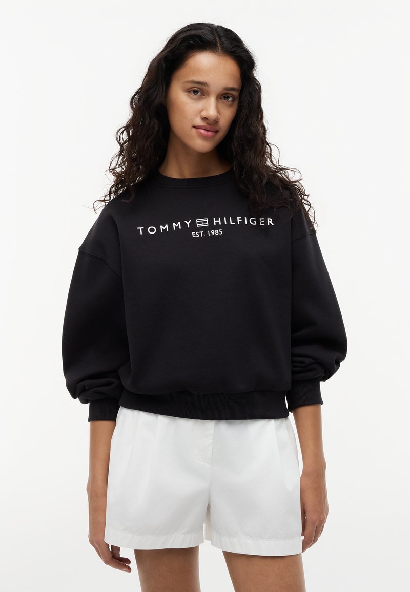 Jeune femme aux longs cheveux bouclés, portant un sweat-shirt noir Tommy Hilfiger et un short blanc, debout devant un fond blanc uni.