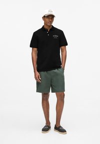 Polo noir, short vert, casquette blanche avec logo et espadrilles noires. Caractéristiques : manches courtes et texte brodé sur le polo.