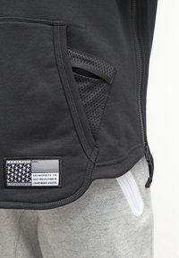 Svart atletisk jacka med dragkedjeficka, med nätdetalj och ett tygmärke som visar ett stjärnmönster och text. Grå träningsoveraller synliga nedanför.