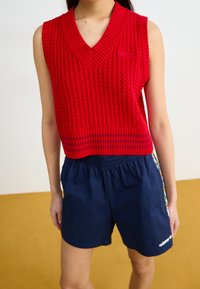 Ärmellose rote Strickweste mit V-Ausschnitt, getragen mit navyblauen, elastischen Sportshorts mit seitlichen Streifen.