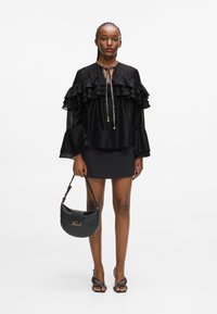 Bluză neagră cu volane și guler legat, asortată cu o fustă mini neagră. Ține în mână o geantă neagră cu formă curbă și logo auriu, purtând sandale cu toc.