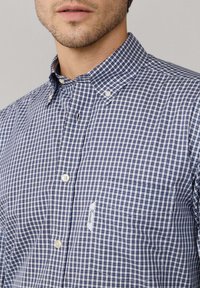 Hombre que lleva una camisa de cuadros azul y blanca, con cuello abotonado y bolsillo en el pecho con etiqueta visible.
