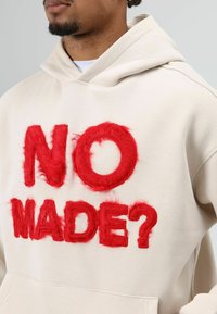 Beige hoodie met grote, zachte rode tekst "NO MADE?". De stof heeft een gladde textuur en een losse pasvorm met een capuchon.