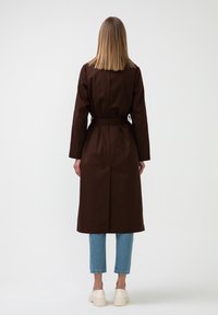 Kvinna med rakt blont hår iklädd en bälte-satt mörkbrun trenchcoat, ljusblå jeans och vita platåskor, stående mot en vit bakgrund.