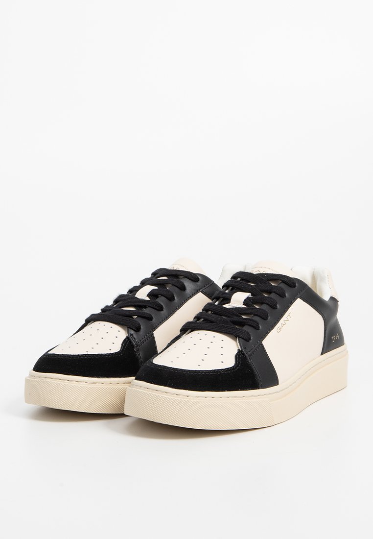 Schwarze und cremefarbene Sneaker mit einem glatten Lederoberteil, perforierter Zehenpartie, Wildlederakzenten und einer dicken strukturierten Sohle. Markenlogo an der Seite.