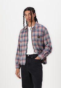 Plaid zip-up shirt in pastel pink and blue, featuring a black logo patch, worn over a white t-shirt with black jeans. Textured fabric.